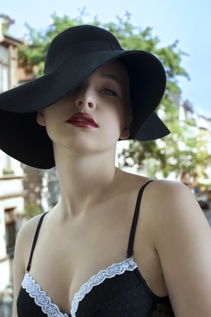 sensual beautiful woman in a fashion shot wearing a black hat and bra outside of the window with old urban background in summer dayの写真素材