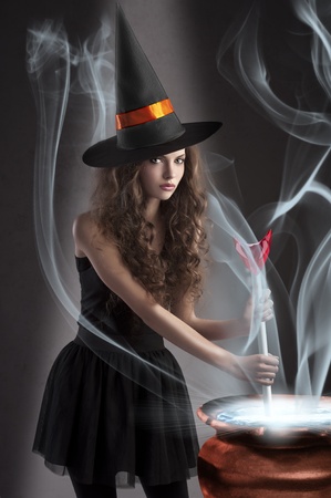 halloween shot of a young and beautiful witch wearing a huge black and orange hat making a poison in a big potの写真素材