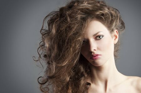 beauty fashion portrait of beautiful young brunette with curly hair flying and creative hairstyle, she is in foreground, looks in to the lens and has tuft on the right.の写真素材