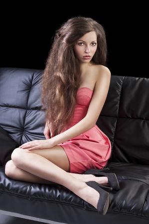 young pretty brunette with hair style wearing an elegant pink dress sitting on a black sofa, she looks in to the lens, her legs are bent on the sofa and she has right hand on the right leg.の写真素材
