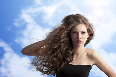 beauty fashion portrait of a very young cute brunette with long curly hair with hairstyle flying in the wind on sky backgroundの写真素材