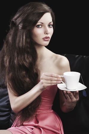 sophisticated young pretty brunette in pink elegant dress sitting on a black sofa and tasting a cup of tea. she looks in to the lens with  three quarters turned faces  and takes a cup of tea with both handsの写真素材