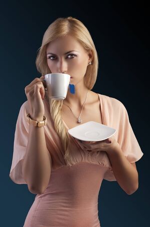 sensual blond girl with hair style in elegant pink dressover holding tea set, drinking tea dark fashion backgroundの写真素材