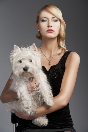 young elegant blond woman wearing black dress with an old fashion hairtyle and necklace jewellery, she is in front of the camera, takes a dog in her arms and looks in to the lensの写真素材