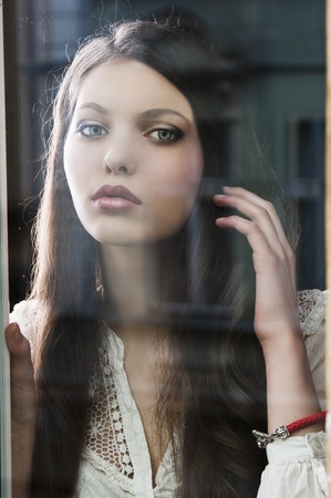 Portrait of a lovely young lady looking through glass window - Indoor in a dark cloudy day, she is in front of the camera and has the left hand near the hairの写真素材