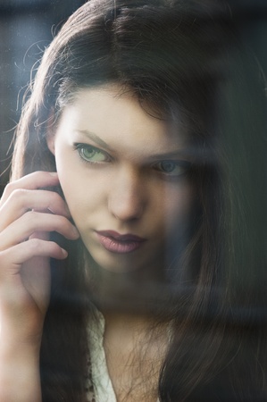 Portrait of a lovely young lady looking through glass window - Indoor in a dark cloudy day, she looks at right and has the right hand near the faceの写真素材