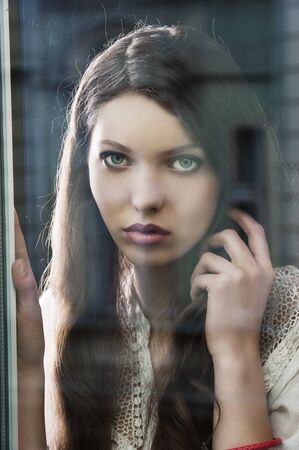 Portrait of a lovely young lady looking through glass window - Indoor in a dark cloudy dayの写真素材