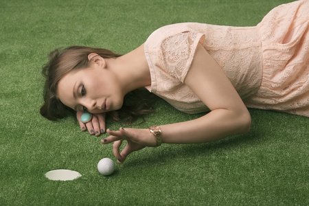 Pretty girl with brown hair lying on grass and playing with golf ball, looks the ball and blows, she is about to hit thatの写真素材