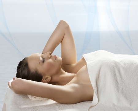 beautiful girl lying on a table with withe towel on her body and under her head, she is in profile, looks in front of her and has both hands near the headの写真素材
