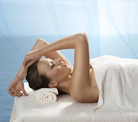 beautiful girl lying on a table with withe towel on her body and under her head, she is in profile with expression of pleasure and with hands near the headの写真素材