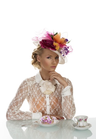 blonde girl with floral hat and vintage shirt behind the table with cake and cup of tea, her face is turned at left and has the left hand under the chinの写真素材