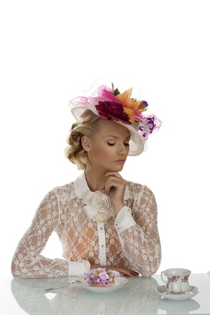 blonde girl with floral hat and vintage shirt behind the table with cake and cup of tea, her eyes are closed and her left hand is under the chinの写真素材