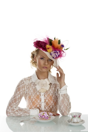 blonde girl with floral hat and vintage shirt behind the table with cake and cup of tea, she is in front of the camera and looks in to the lensの写真素材