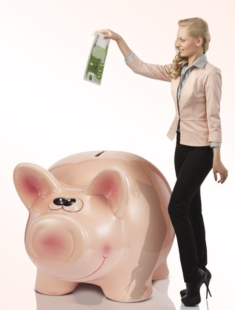 pretty blond girl in elegant dress saving 100 euro in a big pink piggy money bank の写真素材
