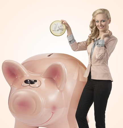 elegant business woman with curly blonde hair-style and elegant suit taking big coin into a big piggy bankの写真素材
