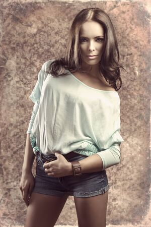 fashion casual woman with long smooth hair wearing pretty vest and denim shorts in sensual pose on grunge backgroundの写真素材