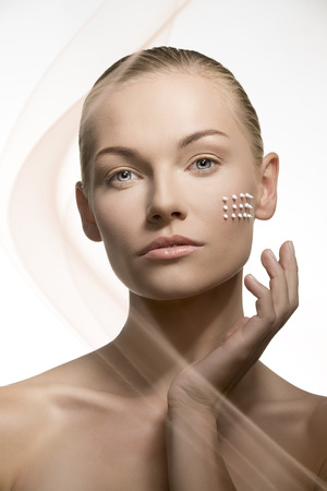 beauty close-up portrait of pretty blonde girl applying white cream on her perfect visage skin, posing with tied back hair and looking in camera の写真素材