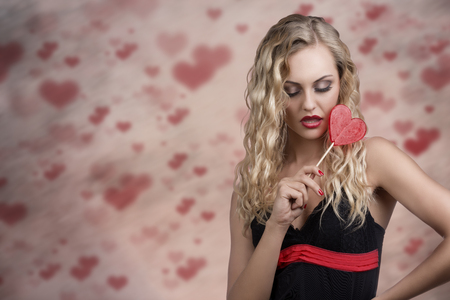 cute young woman with long wavy blonde hair and sexy dress taking heart shaped lollipop in the hand の写真素材