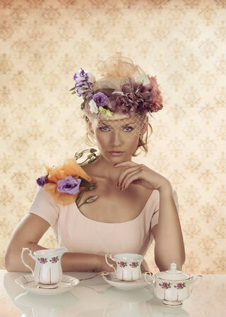 blonde girl with floral decoration and classical tea set on the table, looks in to the lens with left hand under the chinの写真素材
