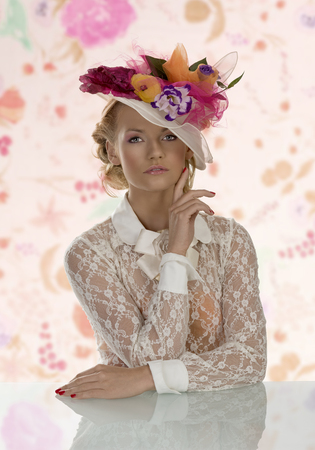 pretty girl behind table with vintage shirt and flowers on the hat, she looks in to the lens with right hand near the faceの写真素材