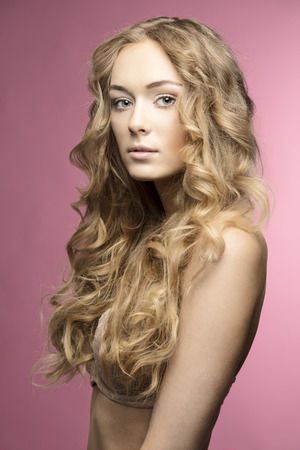 Blonde beautiful young girl with long curly hair in beige lingerie on pink backgroung is looking in cameraの写真素材