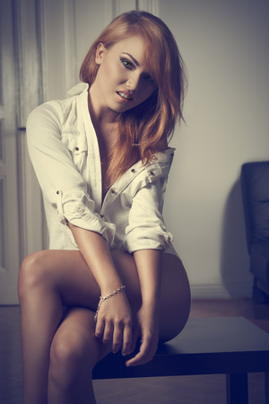 smiling blonde lady in erotic indoor portrait wearing only open shirt and showing her naked legs. Provocative poseの写真素材