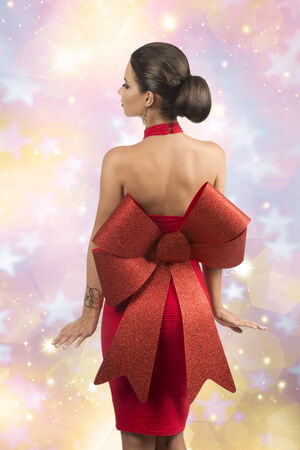 sexy brunette girl with elegant christmas style posing turned on her back with elegant red dress with big glitter bow, naked shoulders and golden jewelleryの写真素材