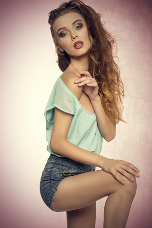fashion portrait of sensual young woman in sexy pose with her perfect body wearing denim shorts, blue shirt and vintage sunglasses on the head. Stylish make-up, charming expressionの写真素材