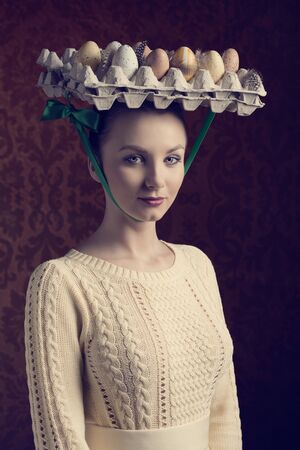 easter portrait of pretty brunette girl with yellow dress and bizarre hat with coloured eggs in carton boxの写真素材
