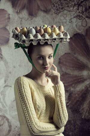 lovely woman with yellow clothes posing in creative easter portrait with bizarre hat with coloured eggs in carton boxの写真素材