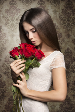Beautiful, lovely, deep in thought woman with brown,straight hair in white dress holding bouquet of red roses.の写真素材