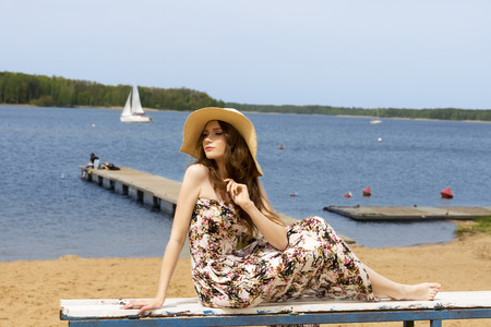 fashion shot of a cute and young woman , wearinf floral dress and summer hat , she is sitting near the lake on the beach.の写真素材