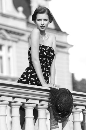 black and white shot ,outside fashion shoot of cute brunette girl with summer elegant style posing on old balcony with short dress and stylish hat in the hand. Looking in camera.の写真素材