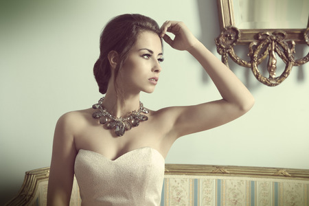 beautiful brunette woman with aristocratic style posing on ancient sofa in indoor fashion shoot with elegant pink dress, shiny necklace and stylish make-up.の写真素材