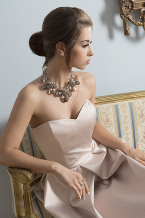 indoor portrait of aristocratic brunette girl sitting on vintage sofa with elegant pink dress, shiny necklace and classic hair-style. Luxury girl in ancient roomの写真素材