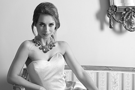 black and white ,fashion indoor portrait of pretty brunette girl with aristocratic style sitting on ancient sofa with classic hair-style, wearing pink elegant dress and shiny necklace. Smiling and looking in cameraの写真素材