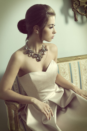 vintage color effect ,indoor portrait of aristocratic brunette girl sitting on vintage sofa with elegant pink dress, shiny necklace and classic hair-style. Luxury girl in ancient roomの写真素材