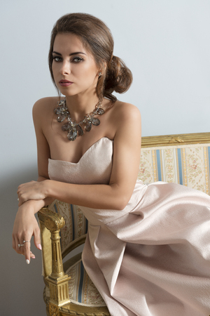 elegant brunette girl with rich style posing with elegant pink dress and precious necklace on old sofa in indoor portraitの写真素材