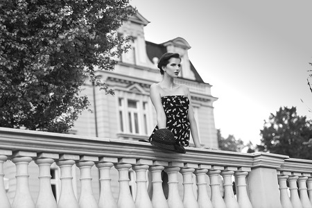 in black and white images very cute woman standing near a parape of and old fashion palace looking in fornt of her. blue elegant dress with hatの写真素材