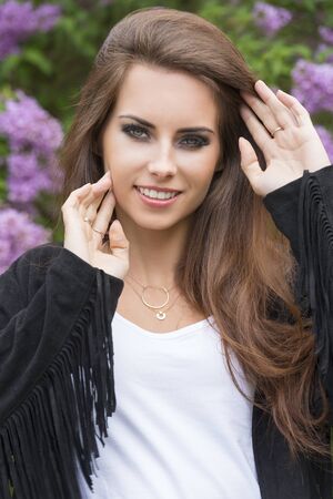 outdoor portrait of happy pretty girl smiling and looking in camera with long hair, stylish black make-up and trendy leather jacket. Flowered green bush on backgroundの写真素材