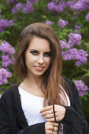 very pretty woman with long natural brown hair, cute make-up and trendy leather jacket posing in outside portrait with flowered green bush on backgroundの写真素材