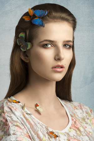 close-up beauty portrait of charming young girl with long brown hair and natural make-up posing with some colorful butterflies on head and shoulderの写真素材