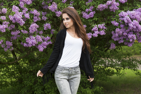 pretty young girl with long natural hair and dark make-up posing in the garden near green flowered bush with fashion jacket and urban jeansの写真素材