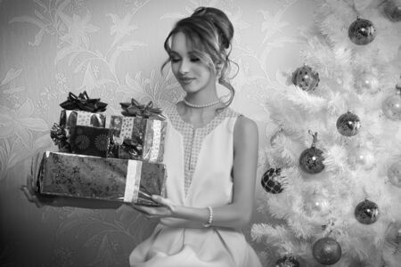 elegant woman in interior xmas portrait sitting near decorated tree with some christmas presents in the hands and looking them black and whiteの写真素材