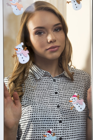 stunning young woman with long hair, freckles, natural make-up and fashion shirt posing indoor trough decorated window in romantic christmas portraitの写真素材