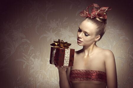 beautiful woman with freckles and artistic creative christmas look. Taking gift box in the hand and posing with golden glitter make-up and red decorated ribbon on hair-style and on breastの写真素材