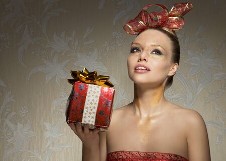 pretty woman with freckles posing with creative golden glitter christmas make-up and gift box in the hand. Red decorated ribbon on hair-style and on breast. Happy expressionの写真素材