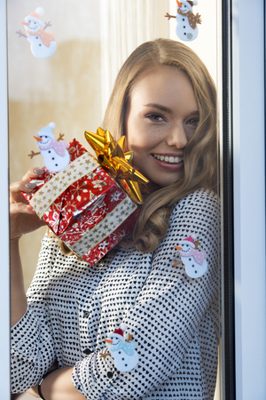 happy smiling  woman with freckles posing in christmas decorated home trough window with natural make-up and fashion shirt. taking gift box in the handの写真素材