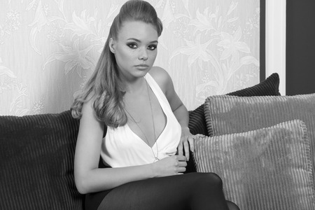 relaxed beautiful blonde girl with cute make-up, long blonde hair and elegant fashion  clothes sitting on sofa and looking in camera , in black and whiteの写真素材