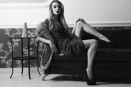stunning woman is sitting in sexy pose on leather sofa in elegant ambient with evening blue dress, heels and fur on shoulder. She is looking in camera and showing her sensual legs .BW shotの写真素材